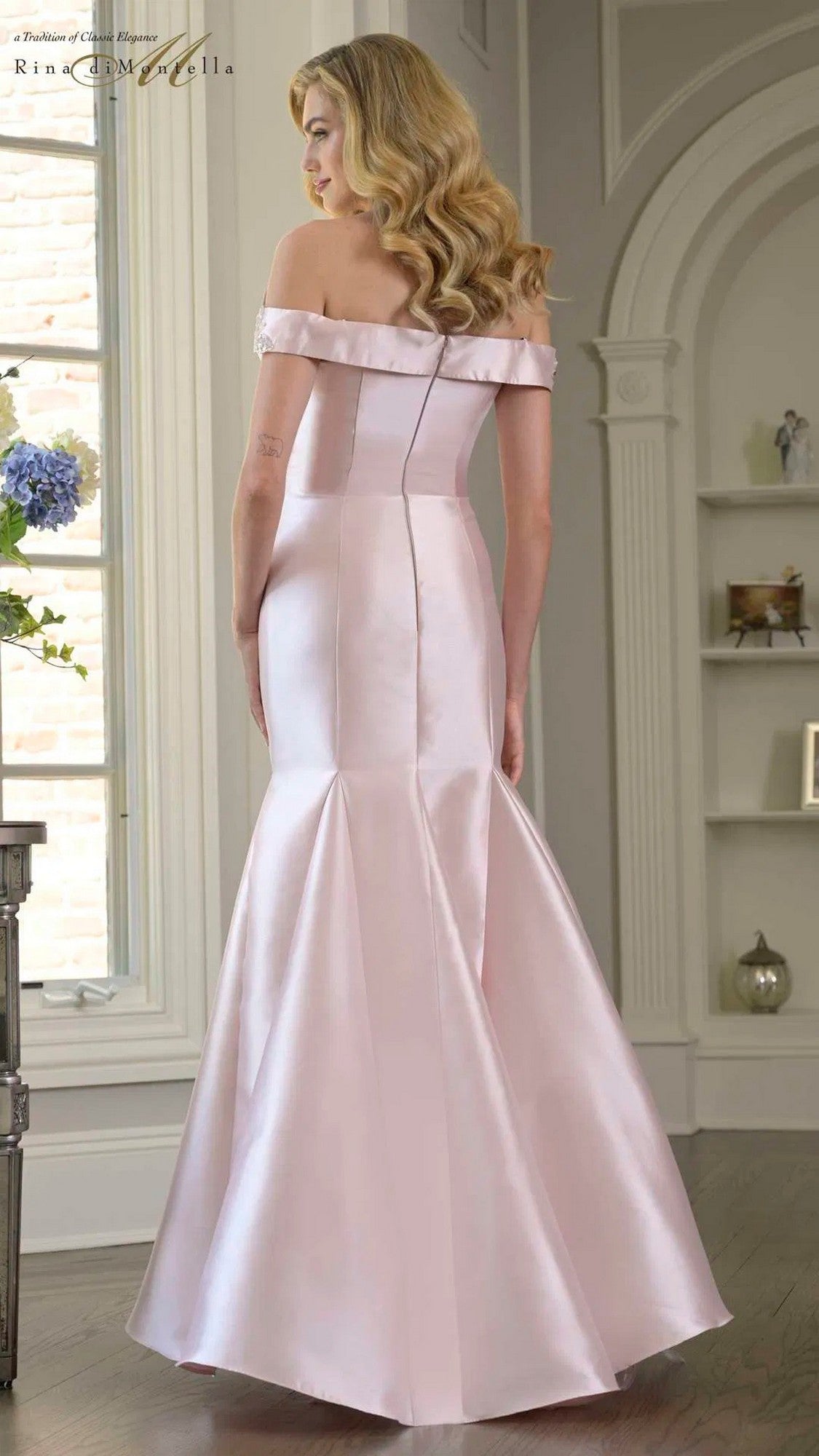 A woman indoors is seen from behind wearing the Rina Di Montella RD3408 long formal dress—a pale pink satin gown featuring an off-the-shoulder neckline, mermaid silhouette, and flared skirt.